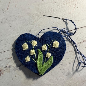 Puede incluir: Un aplique de fieltro azul marino en forma de coraz&oacute;n con un dise&ntilde;o bordado de lirios de los valles. Las flores son de color crema, con hojas y tallos verdes. Una aguja y un hilo azul est&aacute;n unidos, lo que indica un proyecto de manualidades.