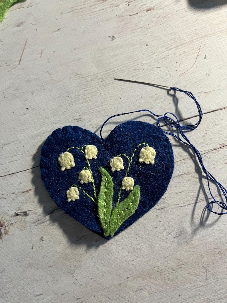 Puede incluir: Un aplique de fieltro azul marino en forma de coraz&oacute;n con un dise&ntilde;o bordado de lirios de los valles. Las flores son de color crema, con hojas y tallos verdes. Una aguja y un hilo azul est&aacute;n unidos, lo que indica un proyecto de manualidades.