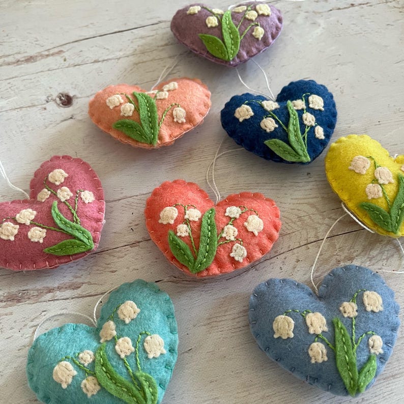 Puede incluir: Una colecci&oacute;n de adornos de fieltro en forma de coraz&oacute;n en varios colores, incluyendo rosa, naranja, azul y amarillo. Cada adorno presenta un dise&ntilde;o bordado de lirios de los valles con flores blancas y hojas verdes. Se adjuntan lazos blancos para colgar.