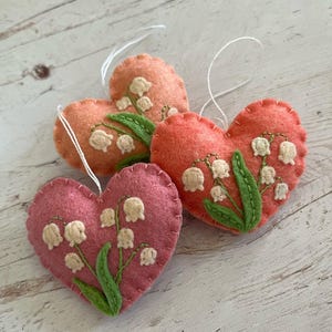 Puede incluir: Tres adornos de fieltro en forma de coraz&oacute;n en tonos rosa y naranja. Cada coraz&oacute;n est&aacute; bordado con flores de lirios de los valles en blanco y verde. Se adjuntan lazos de hilo blanco para colgar. Sobre una superficie de madera clara.