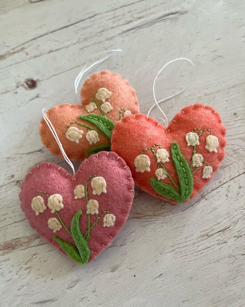 Puede incluir: Tres adornos de fieltro en forma de coraz&oacute;n en tonos rosa y naranja. Cada coraz&oacute;n est&aacute; bordado con flores de lirios de los valles en blanco y verde. Se adjuntan lazos de hilo blanco para colgar. Sobre una superficie de madera clara.