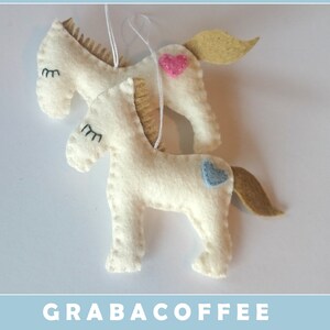 May include: Two white felt horse ornaments with embroidered eyes and a pink and blue heart on each. The horses are hanging from a white string.