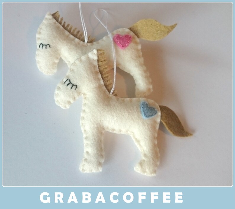 May include: Two white felt horse ornaments with embroidered eyes and a pink and blue heart on each. The horses are hanging from a white string.
