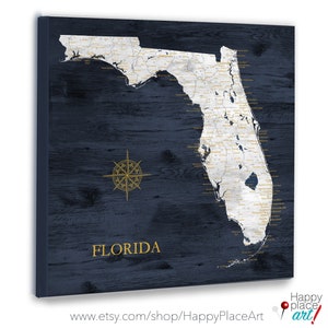 May include: A navy blue wooden map of Florida with gold lettering and a compass rose. The map is ready to be used as a push pin map. The text "FLORIDA" is printed below the map. The text "www.etsy.com/shop/HappyPlaceArt" is printed below the map.