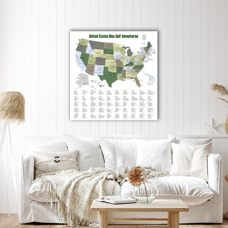 May include: A framed map titled "United States Disc Golf Adventures" hangs on a white wall. The map highlights states in shades of green and gray, with a list of locations below. The room features a white sofa with pillows and a wooden coffee table.