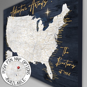 May include: A blue and white map of the United States with gold lettering. The map is on a dark blue wood background. The text on the map says "Adventure Awaits... The Bartons est. 1986". There is a small circle in the lower left corner that says "Push Pin Map Options Available."