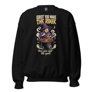 May include: A black long-sleeve sweatshirt featuring a graphic of a witch in a purple hat stirring a large pot of crawfish. The text reads, "FIRST YOU MAKE THE ROUX" in bold yellow letters and "then you cast the spell" in white script.