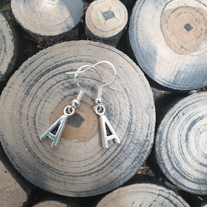 May include: A pair of silver earrings with the letter 'A' hanging from them. The earrings are on a wooden surface.