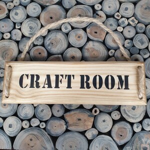 May include: A wooden sign with the text "CRAFT ROOM" in black lettering. The sign is hanging from a rope and is surrounded by a background of wood.