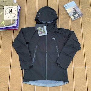 Op de afbeelding: Een zwarte jas met capuchon, voorzien van een volledige ritssluiting aan de voorzijde, lange mouwen met verstelbare manchetten en twee zijzakken met rits. Op de borst staat een grijs logo en er is een Gore-Tex label bevestigd.