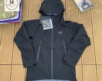 May include: A black hooded jacket featuring a full-length front zipper, long sleeves with adjustable cuffs, and two zippered side pockets. The chest displays a grey logo, and a Gore-Tex tag is attached to the front of the garment.