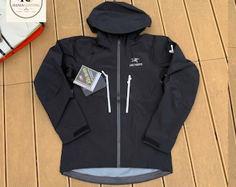 May include: A black Arc'teryx hooded jacket featuring a full-length front zip, two vertical chest pockets with white zips, and a Gore-Tex tag attached. The jacket has long sleeves with adjustable cuffs and a white logo on the chest.