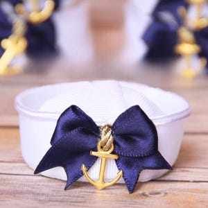 May include: A white sailor hat with a navy blue bow and a gold anchor charm. The bow is made of satin ribbon. The hat is sitting on a wooden surface. Other hats are visible in the background.