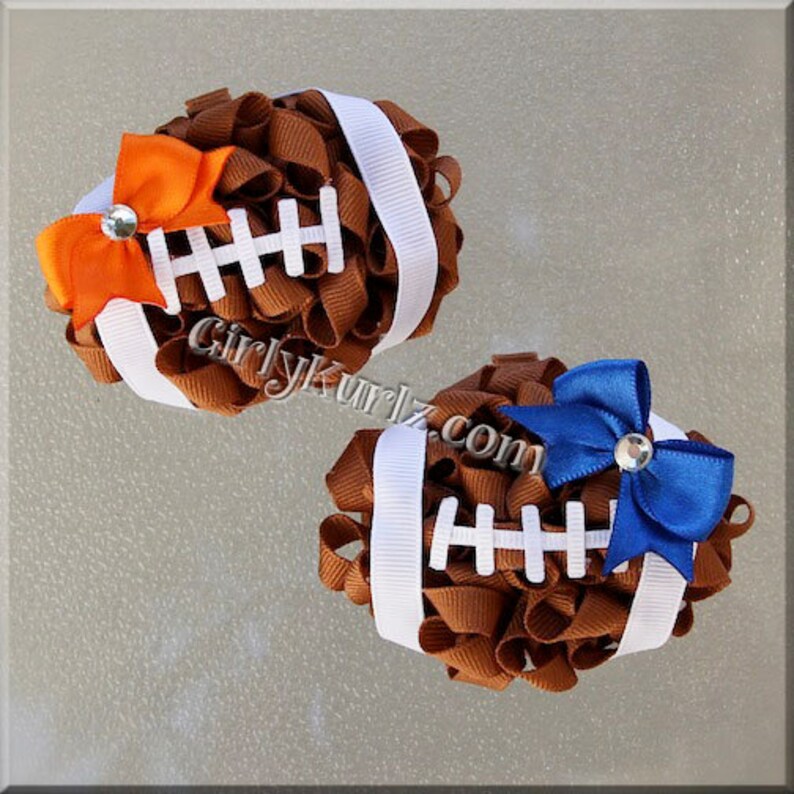 May include: Two decorative football hair bows. The bows are made of brown ribbon with white stripes and accents. One bow has an orange bow with a rhinestone, and the other has a blue bow with a rhinestone. The website "GirlyKurlz.com" is visible.