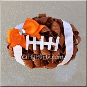 May include: A handmade football-shaped hair bow. The bow is made of brown ribbon with white stripes and white stitching details. An orange bow with a rhinestone center is attached to the top. The text "GirlyKurlz.com" is visible.