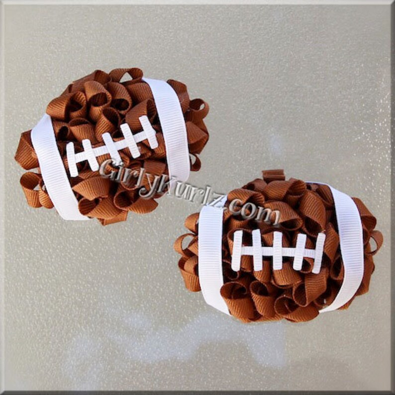 May include: Two brown and white football-shaped hair bows. The bows are made of tightly curled brown ribbon, with white ribbon stripes and white stitching details. The text "GirlyKurlz.com" is visible on the image.