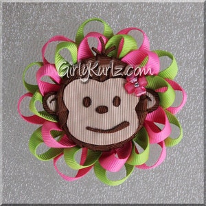 May include: A decorative hair bow featuring a monkey face design. The monkey's face is tan with brown accents, surrounded by layers of pink and green ribbon. The bow includes the text "GirlyKurlz.com".