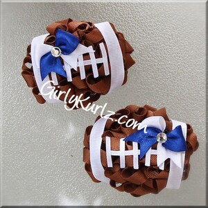 May include: Two brown football-shaped hair accessories with white stripes and a blue bow. Each accessory has a small rhinestone in the center of the bow. The text "GirlyKurlz.com" is visible on the image.