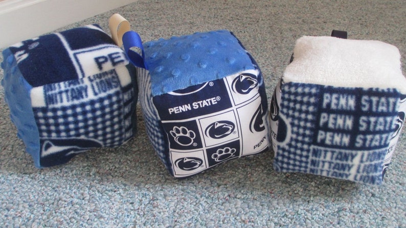 May include: Three blue and white fabric cubes with different patterns. One cube has a blue and white checkered pattern with the text "Penn State" and "Nittany Lions". Another cube has a blue and white checkered pattern with the text "Penn State". The third cube has a blue and white polka dot pattern with the text "Penn State".