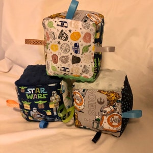 May include: Three soft fabric cubes with different Star Wars themes. The top cube has a white background with various Star Wars characters and symbols. The middle cube has a blue background with the words "Star Wars" and images of Baby Yoda and Mandalorian. The bottom cube has a gray background with the word "Droids" and images of R2-D2 and C-3PO.