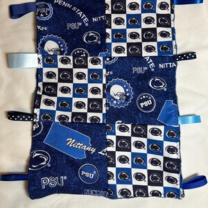 May include: A blue and white baby blanket with a patchwork design featuring the Penn State Nittany Lions logo. The blanket has several blue and white ribbons attached to the corners.