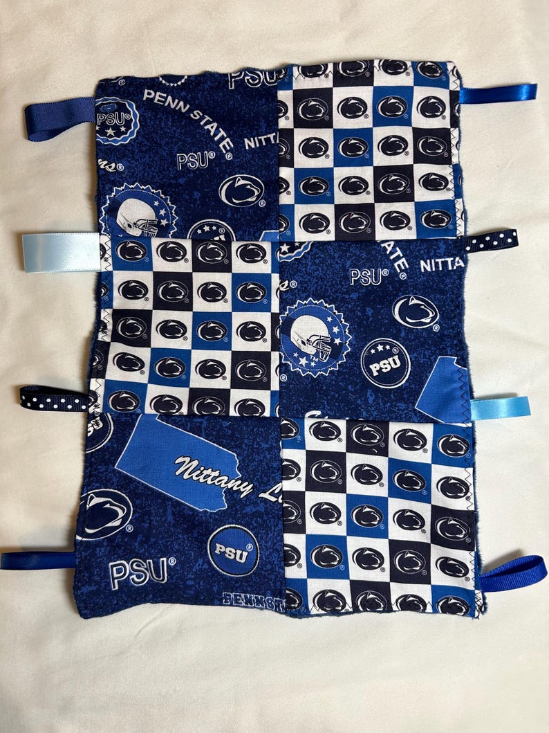 May include: A blue and white baby blanket with a patchwork design featuring the Penn State Nittany Lions logo. The blanket has several blue and white ribbons attached to the corners.