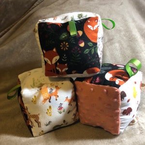 May include: Three soft fabric cubes with different woodland animal prints. The cubes are made of a variety of fabrics, including a soft, textured fabric and a smooth, patterned fabric. The cubes are tied together with green ribbon.