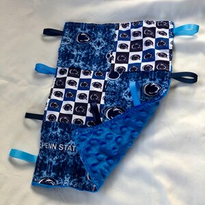 May include: Blue and white baby blanket with a Penn State logo and blue and white checkered pattern. The blanket has blue satin ribbons attached to the corners.