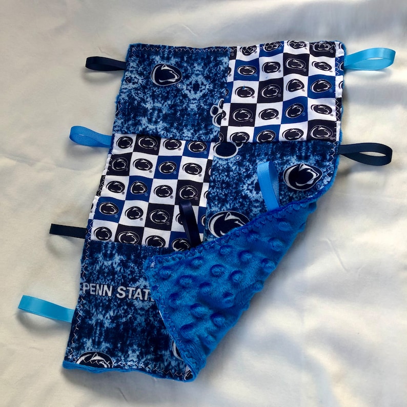 May include: Blue and white baby blanket with a Penn State logo and blue and white checkered pattern. The blanket has blue satin ribbons attached to the corners.