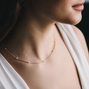 May include: A delicate gold necklace featuring small, evenly spaced pearls. The necklace is draped around a person's neck, against a white dress. The gold chain is thin and elegant, creating a minimalist and refined aesthetic.