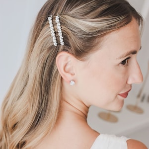 May include: A woman with long blonde hair wearing a white dress and a pair of silver earrings. She has two silver hair clips with clear rhinestones in her hair.