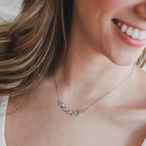 May include: A silver necklace with a delicate leaf design. The necklace is worn by a person with long brown hair and a white shirt.