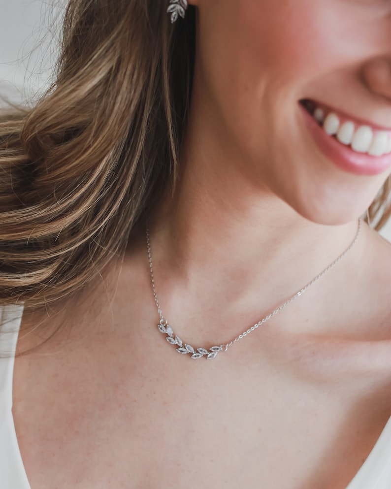May include: A silver necklace with a delicate leaf design. The necklace is worn by a person with long brown hair and a white shirt.