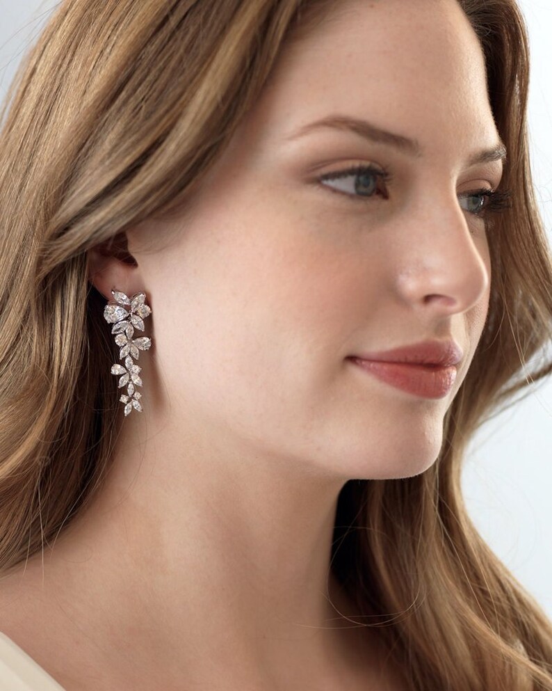 May include: A pair of long, silver dangle earrings with a floral design. The earrings are made of clear crystals and have a delicate, feminine look.