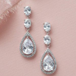 May include: A pair of silver teardrop earrings with clear crystal stones. The earrings have a pear-shaped stone in the center, surrounded by a halo of smaller stones. The earrings are suspended from a silver chain with two smaller oval-shaped stones.