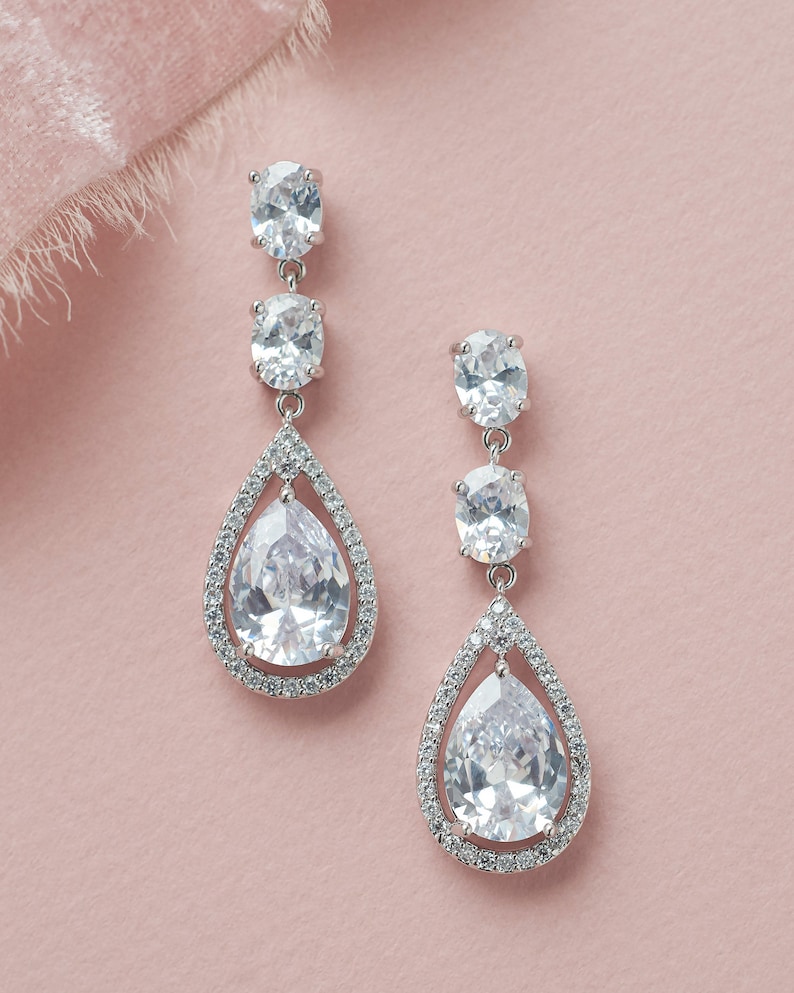 May include: A pair of silver teardrop earrings with clear crystal stones. The earrings have a pear-shaped stone in the center, surrounded by a halo of smaller stones. The earrings are suspended from a silver chain with two smaller oval-shaped stones.