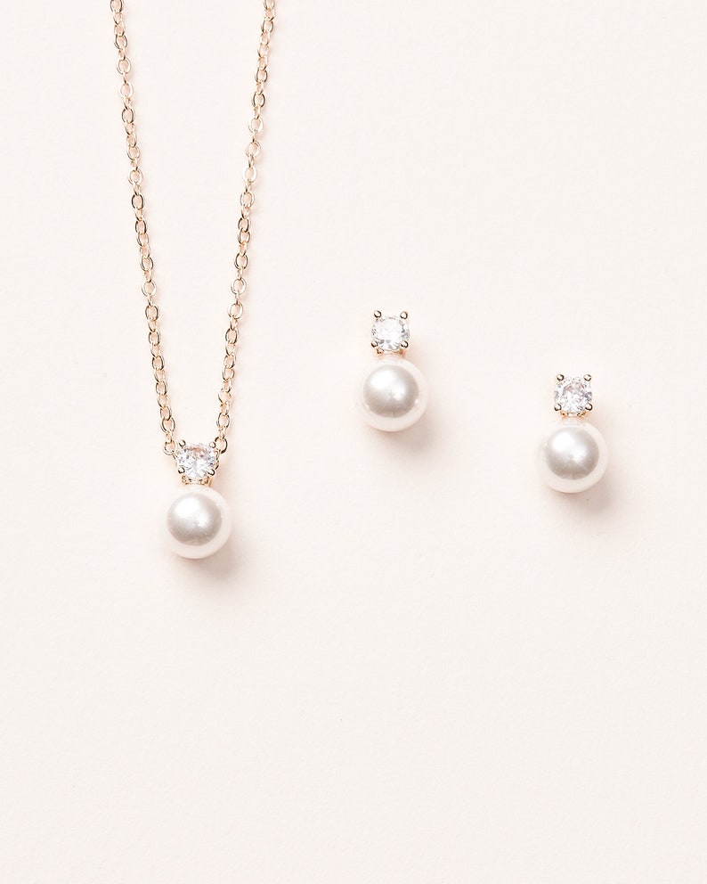 May include: A jewellery set consisting of a necklace and two stud earrings. Each piece features a round white pearl suspended beneath a single clear, faceted round gemstone set in a rose gold-toned metal setting on a delicate matching chain.