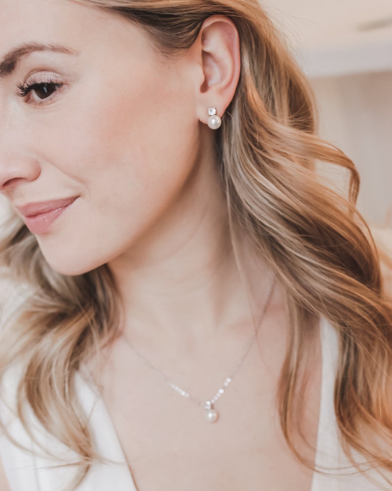 May include: A silver-toned necklace featuring a single white pearl pendant suspended from a delicate chain, paired with matching stud earrings that each consist of a small clear stone set above a round white pearl.