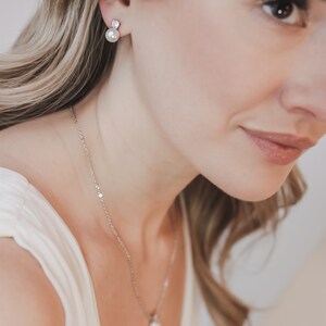 May include: A silver-toned necklace featuring a small, round white pearl pendant on a delicate chain, paired with matching stud earrings. Each earring consists of a single white pearl suspended beneath a small, clear, faceted stone setting.
