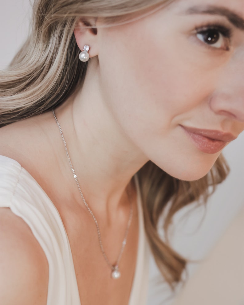 May include: A silver-toned necklace featuring a small, round white pearl pendant on a delicate chain, paired with matching stud earrings. Each earring consists of a single white pearl suspended beneath a small, clear, faceted stone setting.