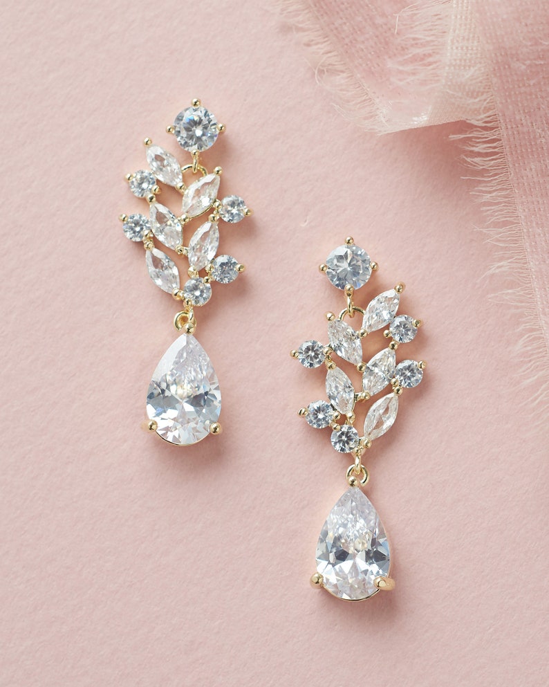 Puede incluir: Un par de pendientes colgantes de oro con gotas de cristal transparente en forma de pera y un racimo de cristales transparentes m&aacute;s peque&ntilde;os en un dise&ntilde;o de hoja.