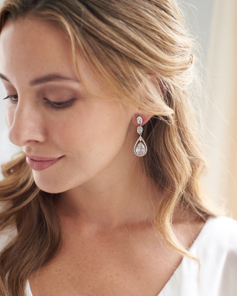May include: A pair of silver teardrop earrings with clear crystal stones. The earrings have a pear-shaped stone in the center, surrounded by smaller stones.