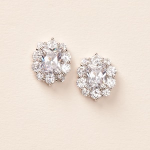May include: A pair of silver-toned stud earrings. Each earring features a large, square, clear gemstone surrounded by smaller, round, clear gemstones. The earrings are set against a cream-colored background.