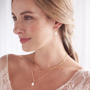 Pu&ograve; includere: Una donna indossa una camicetta di pizzo bianco con una collana a catena d'oro e un ciondolo di perla. Indossa anche orecchini d'oro con gocce di perle.