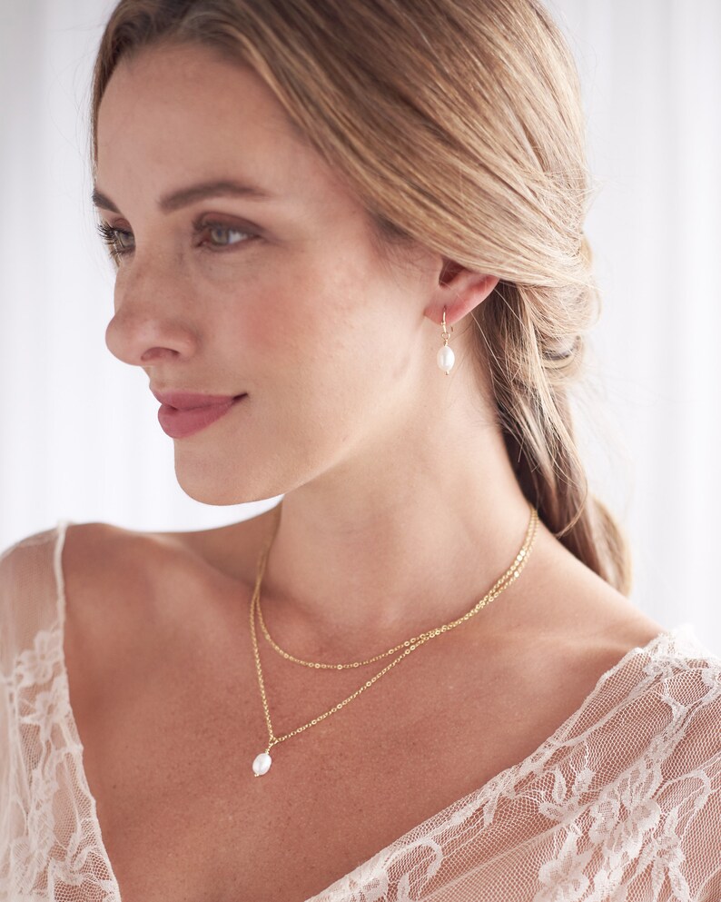 Pu&ograve; includere: Una donna indossa una camicetta di pizzo bianco con una collana a catena d'oro e un ciondolo di perla. Indossa anche orecchini d'oro con gocce di perle.