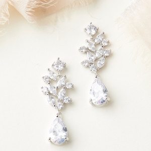Puede incluir: Un par de pendientes colgantes de plata con piedras de cristal transparentes en forma de pera y piedras de cristal transparentes m&aacute;s peque&ntilde;as dispuestas en forma de hoja.