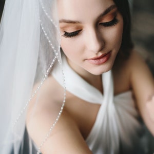 K&ouml;nnte beinhalten: Eine Braut in einem wei&szlig;en Brautkleid mit einem Schleier, der mit einer Perlenborte verziert ist. Der Schleier ist &uuml;ber ihren Kopf und ihre Schultern gelegt, und sie schaut nach unten.