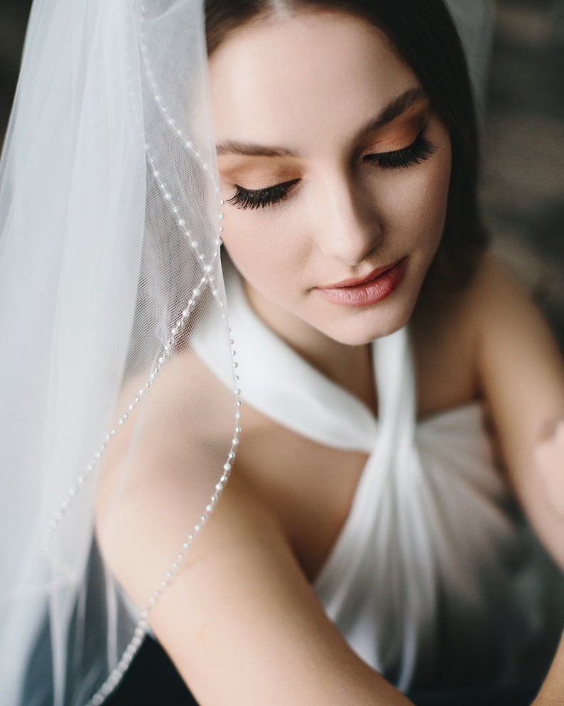 K&ouml;nnte beinhalten: Eine Braut in einem wei&szlig;en Brautkleid mit einem Schleier, der mit einer Perlenborte verziert ist. Der Schleier ist &uuml;ber ihren Kopf und ihre Schultern gelegt, und sie schaut nach unten.