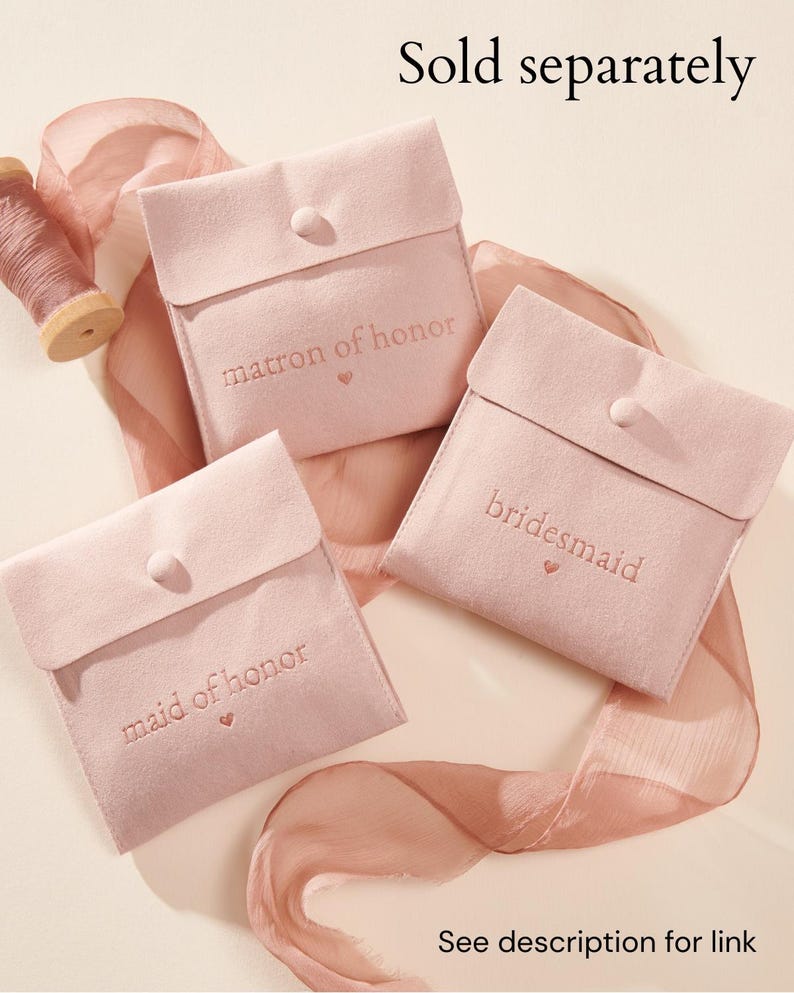 May include: Three light pink square pouches with button closures, each embroidered with the text matron of honour, bridesmaid, or maid of honour above a small heart. A spool of matching pink ribbon is positioned near the pouches.