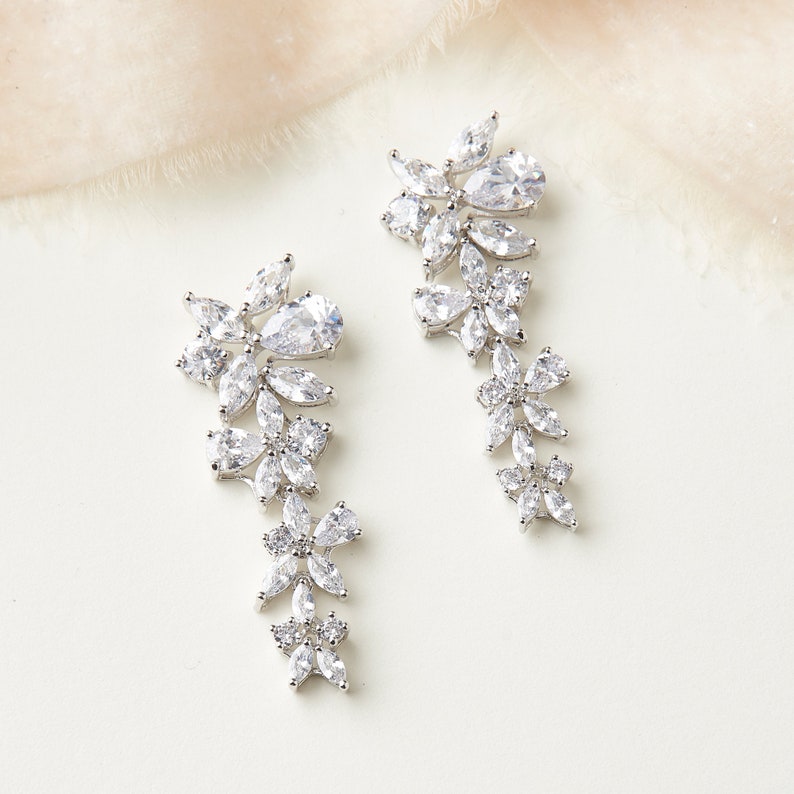 May include: A pair of silver dangle earrings with a floral design featuring clear cubic zirconia stones.